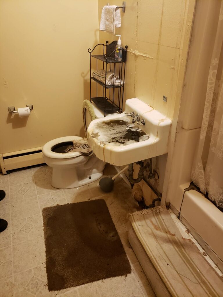 A dirty bathroom showing an overflowing sink and toilet with dark, foamy water