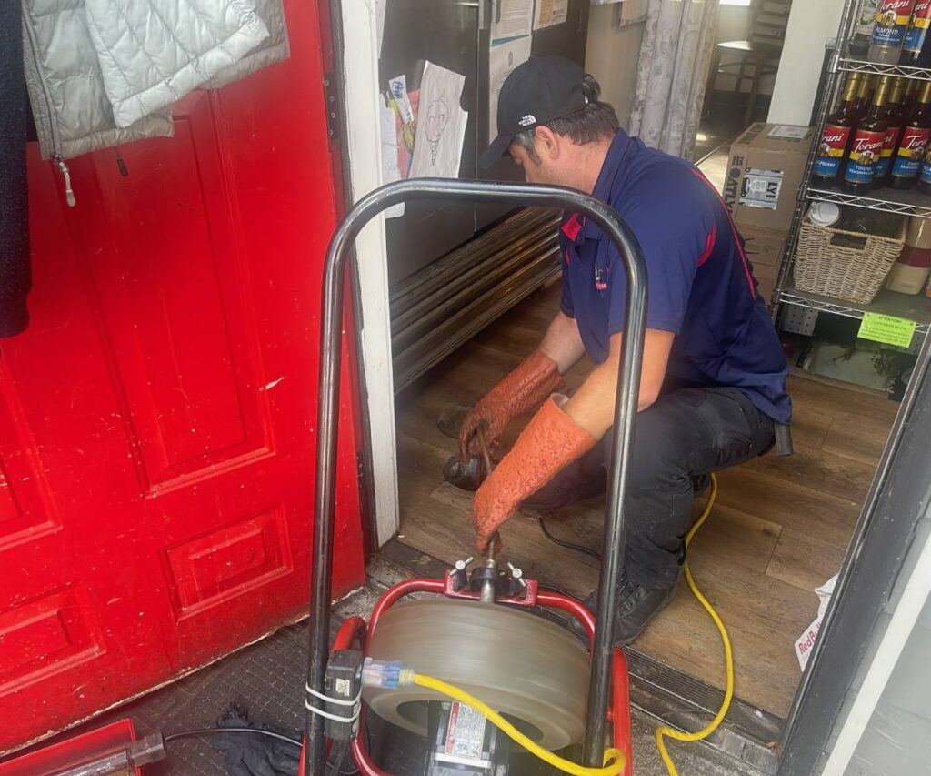 A plumber operating a drain snake machine to clear a floor drain inside a building