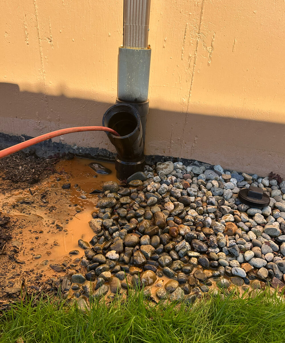 A red hydro-jetting hose extending into a downspout pipe next to a building, with muddy water pooling around decorative rocks