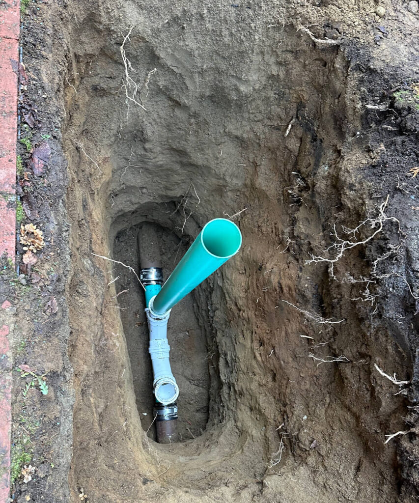 A deep trench exposing new green and grey plumbing pipes installed to connect with older existing lines