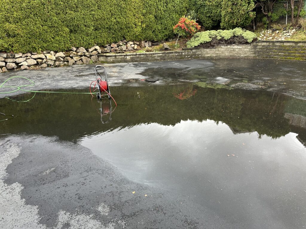 A large, dark puddle covers an asphalt surface, reflecting the cloudy sky and green hedges