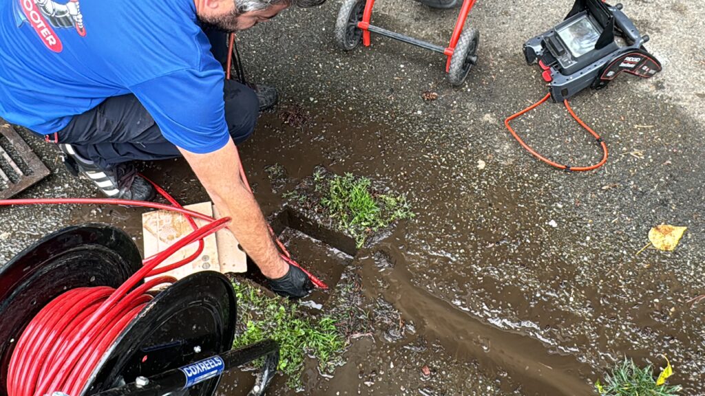 A Knight Rooter technician feeding a red drain cable into a muddy outdoor sewer drain opening